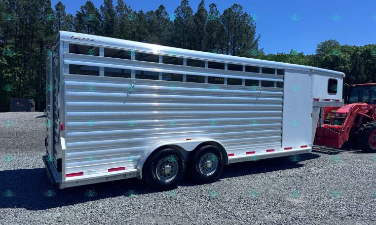 Silver aluminum livestock trailer with three axles parked on a gravel lot, trees in the background and a blue sky, watermark visible along the bottom edge.]