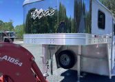 Polished aluminum horse trailer with an Exiss logo, reflecting trees and blue sky, hitched to a red tractor attachment in a gravel lot.