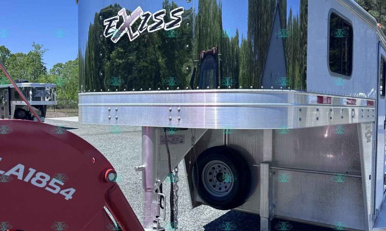 Polished aluminum horse trailer with an Exiss logo, reflecting trees and blue sky, hitched to a red tractor attachment in a gravel lot.