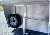Interior of aluminum trailer showing a mounted spare tire, support leg, and metal wall panels in a gravel lot.