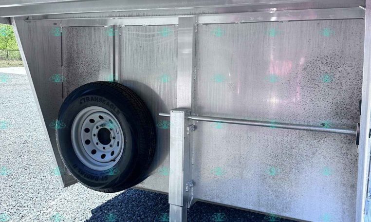 Interior of aluminum trailer showing a mounted spare tire, support leg, and metal wall panels in a gravel lot.