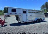 White aluminum livestock trailer with slatted side panels and a small rectangular window, parked on a gravel lot with trees in the background; watermark reads CarolinaTrailersales.com at bottom.