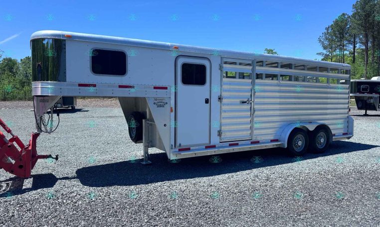 White aluminum livestock trailer with slatted side panels and a small rectangular window, parked on a gravel lot with trees in the background; watermark reads CarolinaTrailersales.com at bottom.