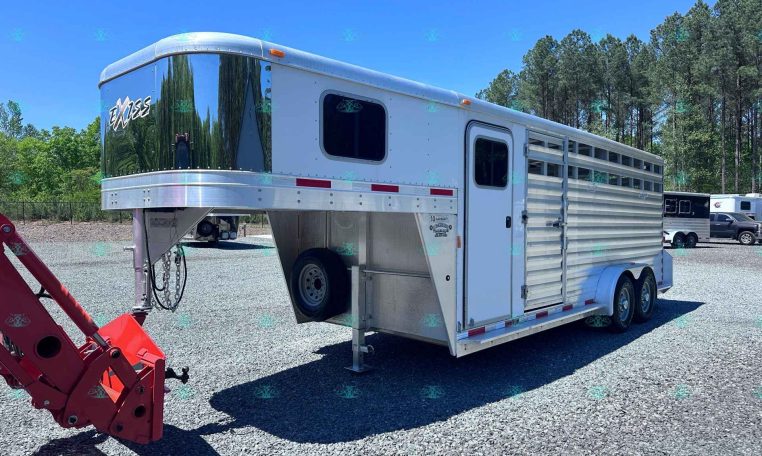 White gooseneck horse trailer with a chrome front, parked on a gravel lot with trees in the background.