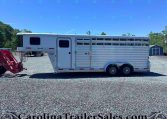 White, aluminum horse trailer with multiple windows and a gooseneck hitch on a gravel lot under a clear blue sky.