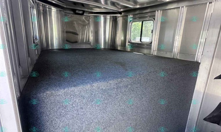 Interior of an empty aluminum cargo trailer with metal walls, ceiling rails, and a blue-gray carpeted floor; side window visible.