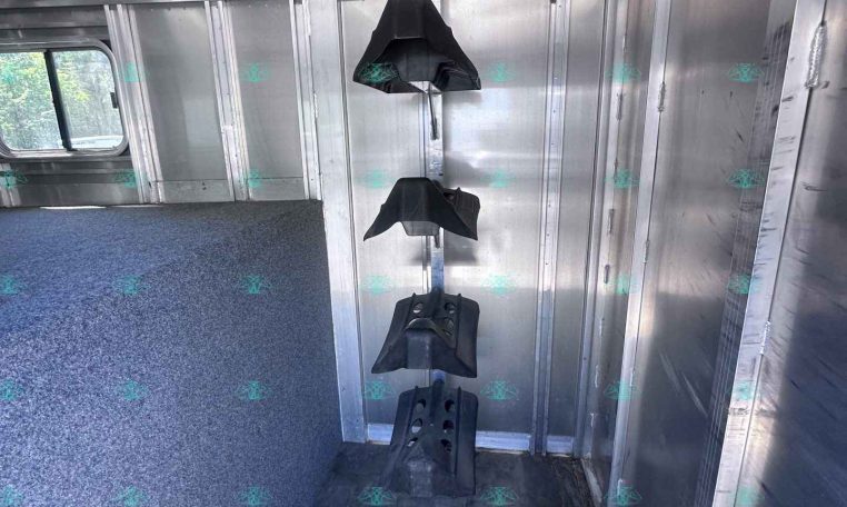 Interior of an aluminum horse trailer with vertical saddle racks along the wall and a blue speckled floor, seen from the doorway.