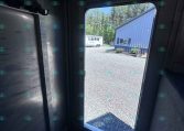 Interior of a metal trailer with an open door, revealing a gravel yard and a blue metal building outside. A white trailer is visible in the distance among trees.