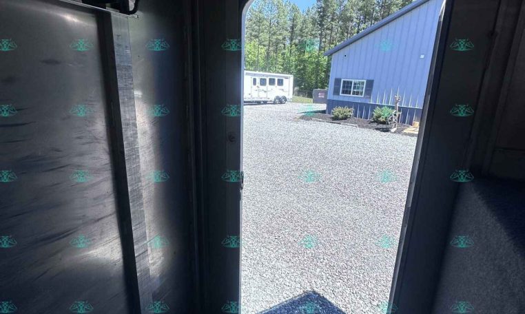 Interior of a metal trailer with an open door, revealing a gravel yard and a blue metal building outside. A white trailer is visible in the distance among trees.
