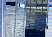 Rear view of a silver aluminum trailer with the ramp door open, showing ventilated slatted sides and a dark interior.