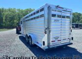 Silver aluminum livestock trailer parked on a gravel lot with green trees and a blue sky in the background, branded EXISS by Universal Trailer.
