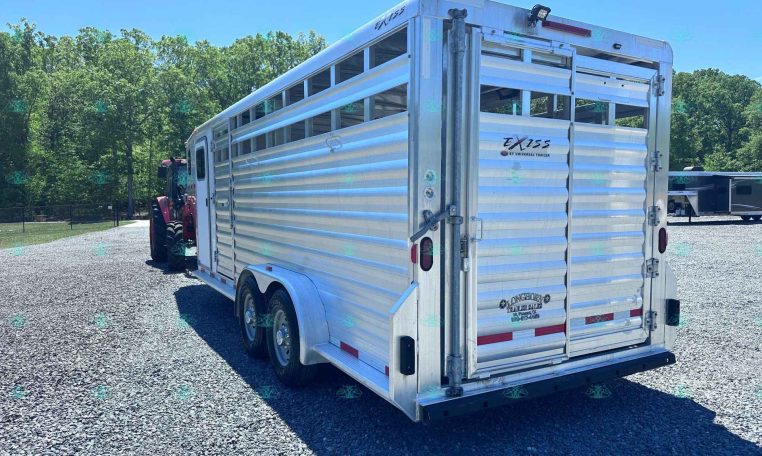 Silver aluminum livestock trailer parked on a gravel lot with green trees and a blue sky in the background, branded EXISS by Universal Trailer.