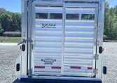 Rear view of a silver EXISS livestock trailer on a gravel lot, blue sky and trees in the background.