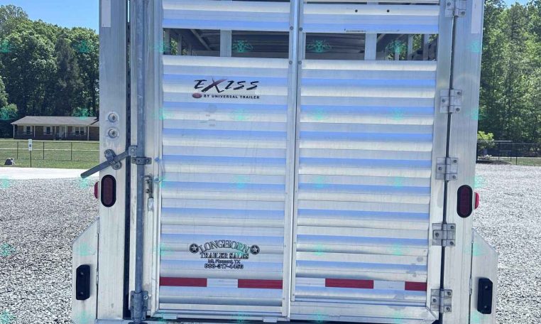 Rear view of a silver EXISS livestock trailer on a gravel lot, blue sky and trees in the background.