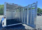 Open aluminum livestock trailer with rear doors swung wide, interior empty, on a gravel lot under a blue sky.
