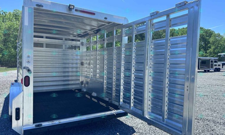 Open aluminum livestock trailer with rear doors swung wide, interior empty, on a gravel lot under a blue sky.