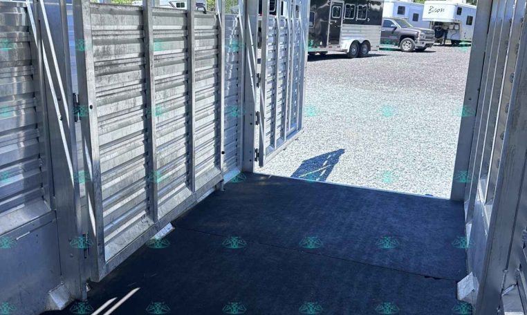 Interior of a metal trailer with ribbed aluminum walls, a black non-slip floor, and an open rear doorway; other trailers are visible outside in a gravel lot.