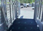 Inside a metal livestock trailer, view toward an open rear with several horse trailers and trucks in a gravel lot beyond.