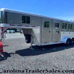 Large gray gooseneck horse trailer with living quarters, attached to a red tow hitch, parked on a gravel lot with trees in the background.