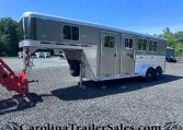 Large gray gooseneck horse trailer with living quarters, attached to a red tow hitch, parked on a gravel lot with trees in the background.