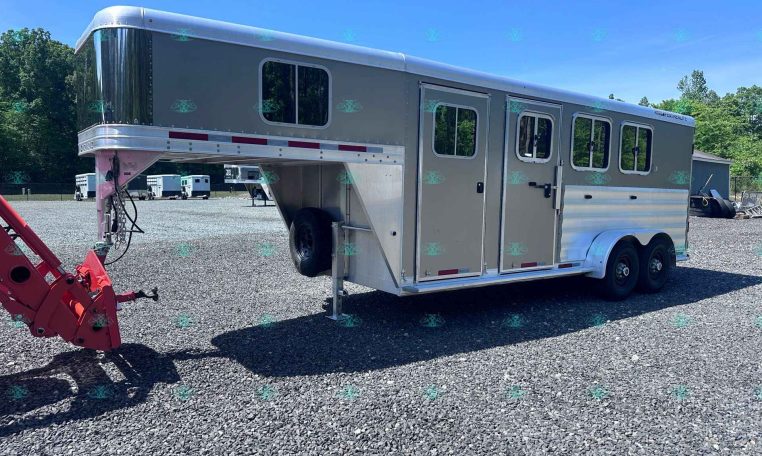 Large gray gooseneck horse trailer with living quarters, attached to a red tow hitch, parked on a gravel lot with trees in the background.