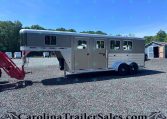 Gray gooseneck horse trailer with side windows parked on a gravel lot, with trees and a blue shed in the background.