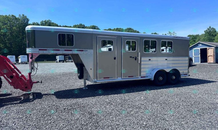 Gray gooseneck horse trailer with side windows parked on a gravel lot, with trees and a blue shed in the background.
