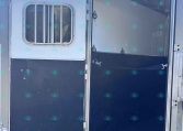 Open side door of a silver horse trailer; interior stalls visible through the doorway with a windowed side panel