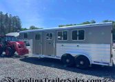 Side view of a large silver Featherlite horse trailer with multiple windows, parked on a gravel lot; a red tractor sits at its front and trees line the background under a blue sky.