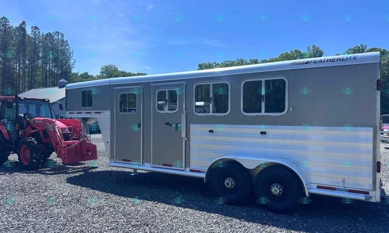 Side view of a large silver Featherlite horse trailer with multiple windows, parked on a gravel lot; a red tractor sits at its front and trees line the background under a blue sky.