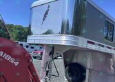 Shiny aluminum travel trailer nose with riveted trim, parked in a gravel lot under a blue sky.