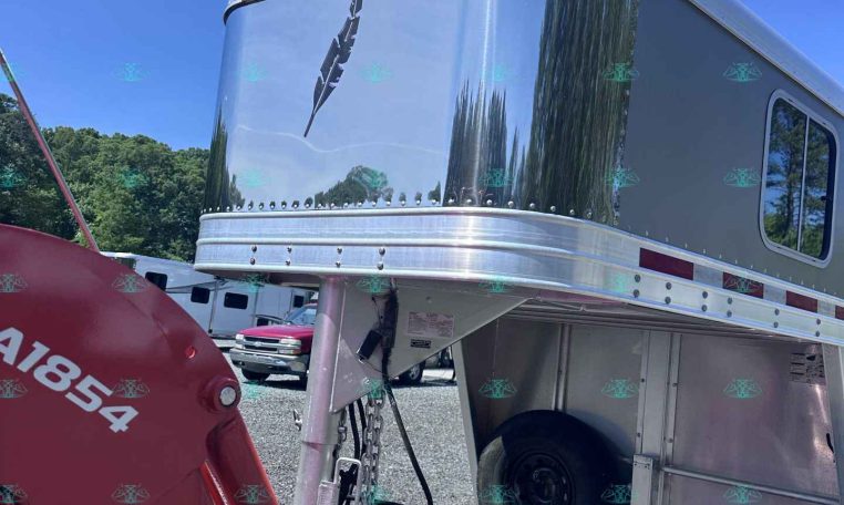 Shiny aluminum travel trailer nose with riveted trim, parked in a gravel lot under a blue sky.