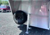 Rear side of a metal trailer with a mounted spare tire on the wheel well and a stabilizing jack leg on a gravel surface