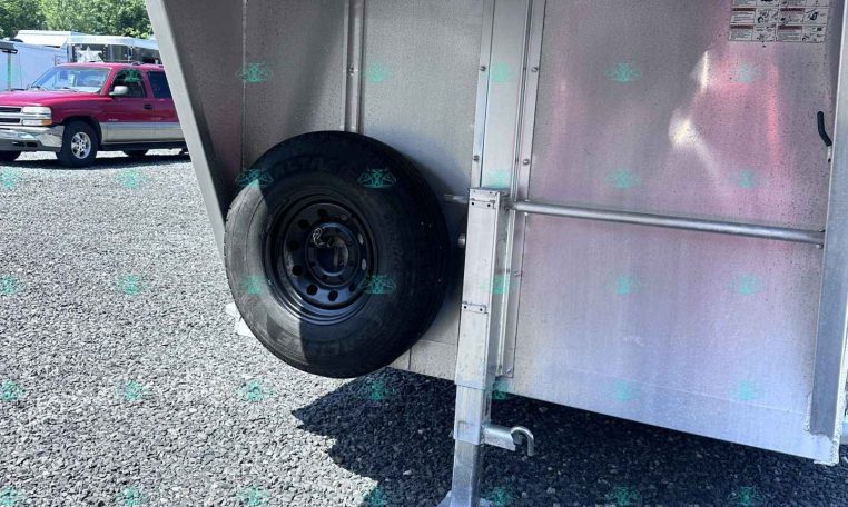 Rear side of a metal trailer with a mounted spare tire on the wheel well and a stabilizing jack leg on a gravel surface