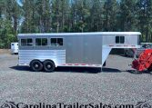 Silver Featherlite horse trailer with multiple side windows on a gravel lot, front section raised for living quarters, with trees in the background.