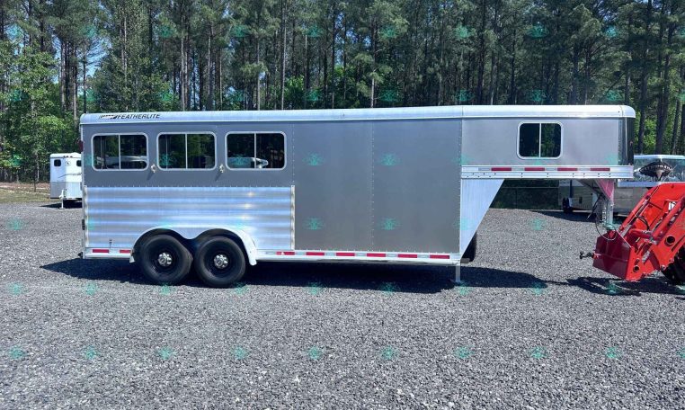 Silver Featherlite horse trailer with multiple side windows on a gravel lot, front section raised for living quarters, with trees in the background.