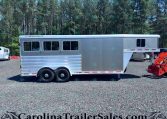 Silver Featherlite horse trailer with rounded windows and tandem axles parked on a gravel lot, with a forest backdrop of tall trees.