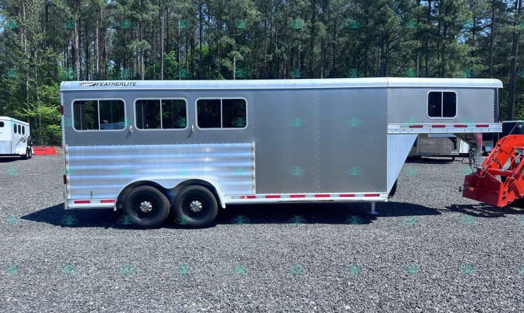 Silver Featherlite horse trailer with rounded windows and tandem axles parked on a gravel lot, with a forest backdrop of tall trees.