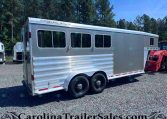 Silver enclosed trailer with three side windows and dual wheels on a gravel lot, trees in the background.