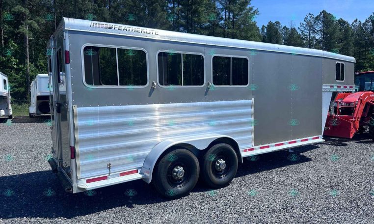 Silver enclosed trailer with three side windows and dual wheels on a gravel lot, trees in the background.