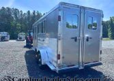 Rear corner of a silver horse trailer on a gravel lot with trees in the background.