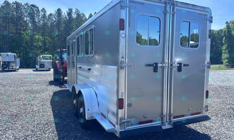 Rear corner of a silver horse trailer on a gravel lot with trees in the background.