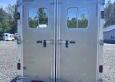 Rear view of a silver aluminum trailer with double doors, small windows, and latches on a gravel lot with trees in the background
