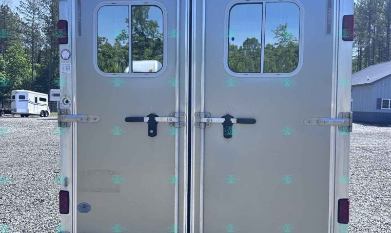 Rear view of a silver aluminum trailer with double doors, small windows, and latches on a gravel lot with trees in the background