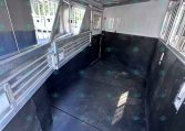 Interior of a shiny aluminum horse trailer with side dividers, windows along the right, and a black rubber floor, viewed from the rear toward the front.