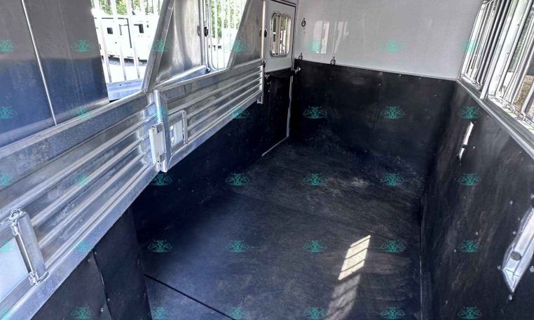 Interior of a shiny aluminum horse trailer with side dividers, windows along the right, and a black rubber floor, viewed from the rear toward the front.