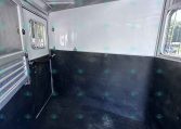 Interior of an aluminum trailer: silver walls, a small windowed door on the left, dark lower panels, and a rubberized floor. Light from an open side window hints at outside scenery.