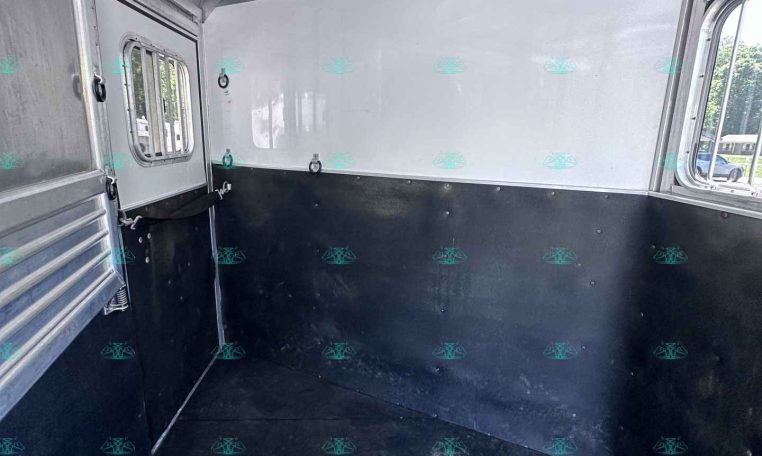 Interior of an aluminum trailer: silver walls, a small windowed door on the left, dark lower panels, and a rubberized floor. Light from an open side window hints at outside scenery.