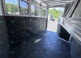 Interior of a metal horse trailer with padded black walls, row of barred windows along the left, and an open rear ramp to a gravel yard with trees outside