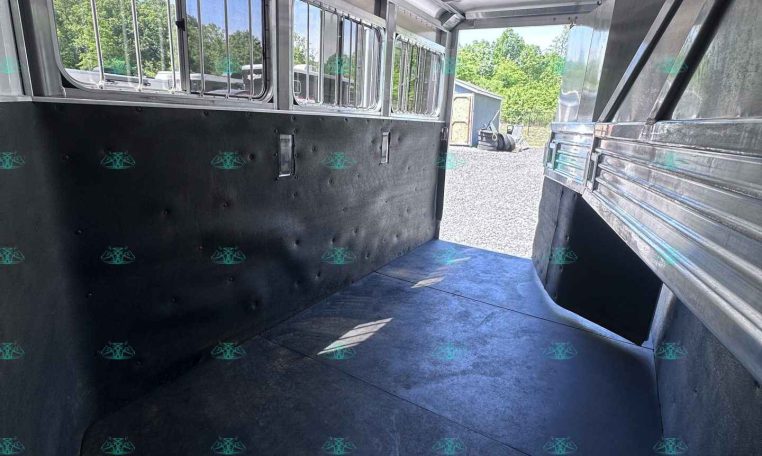 Interior of a metal horse trailer with padded black walls, row of barred windows along the left, and an open rear ramp to a gravel yard with trees outside